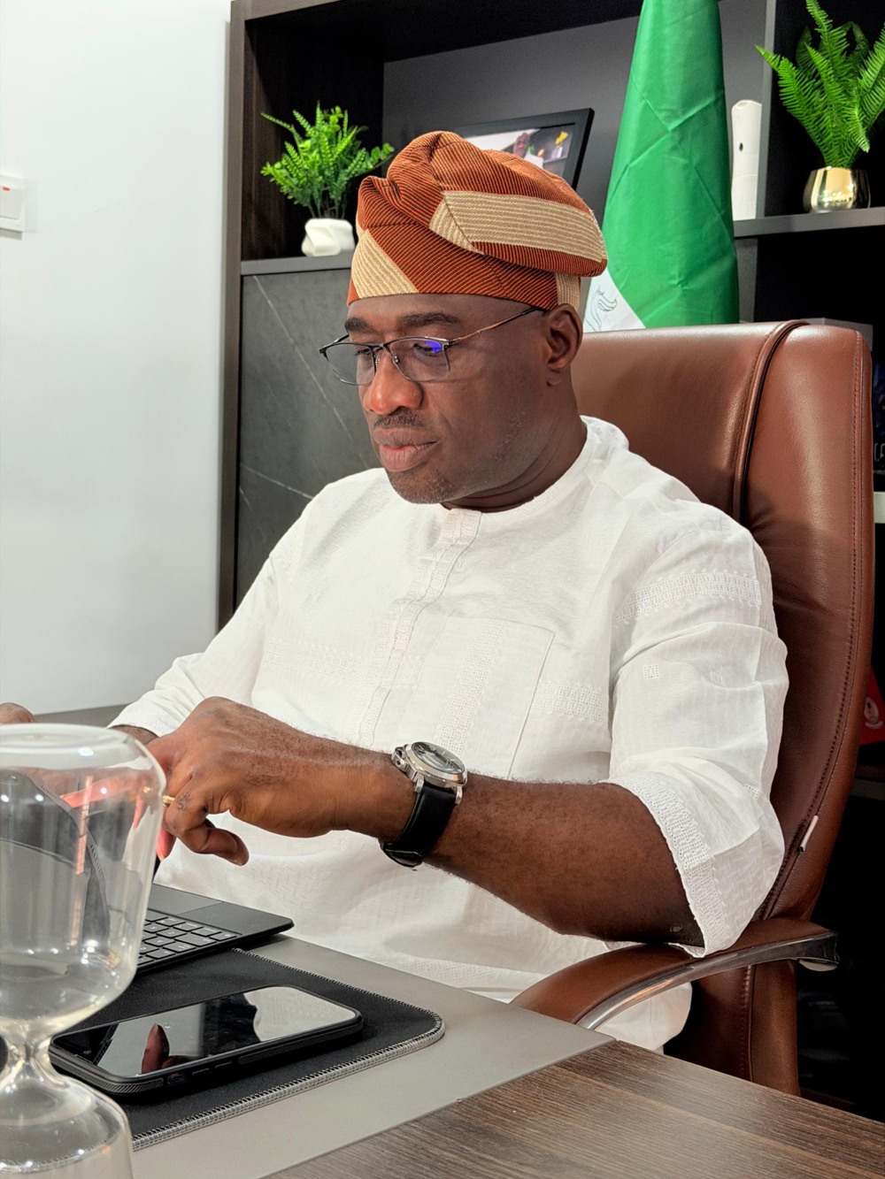 Prince Idowu Ilara wearing a white traditional outfit and an orange patterned cap, seated at his desk.